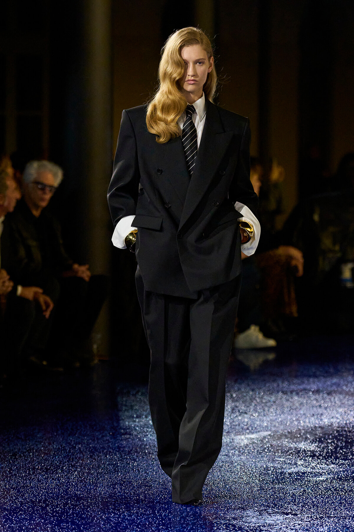 Female model walks a fashion runway in an oversized black double-breasted suit with white cuffs and a striped tie on a glittery blue catwalk.