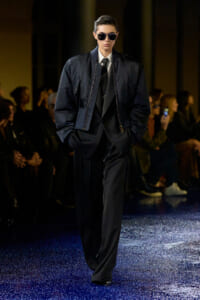 Male model walking a runway in a dark suit with an oversized navy bomber jacket and sunglasses, hands in pockets, spotlighted on a glittering blue runway.