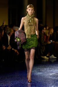 Model walks a fashion runway in a sheer lace gold top and layered green mini skirt, carrying a colorful patterned coat or accessory on her arm.