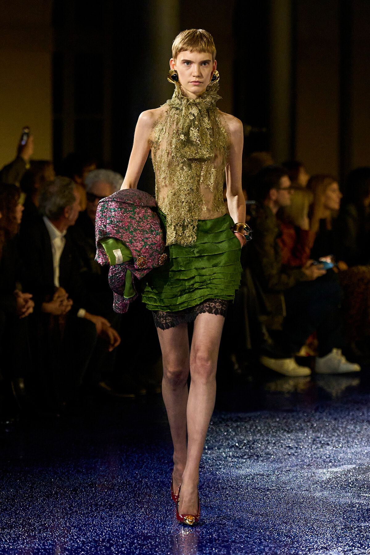 Model walks a fashion runway in a sheer lace gold top and layered green mini skirt, carrying a colorful patterned coat or accessory on her arm.