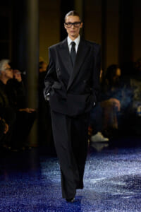 Model on a runway wearing an oversized black suit with a white shirt and black tie, hands in pockets, glasses on.