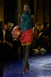 Model walks the runway in a teal lace high-neck top and a layered red mini skirt with gold lace detail, holding a sparkly scarf; gold jewelry and red heels visible.