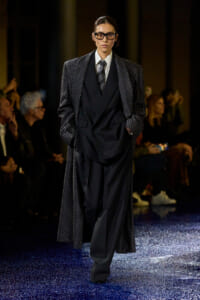 Model on a fashion runway wearing an oversized black suit with a long polka-dot coat and dark glasses.