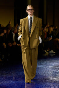 Model walks a fashion runway in an oversized tan double-breasted suit, white shirt, striped tie, and black-framed glasses amid a dark audience backdrop.