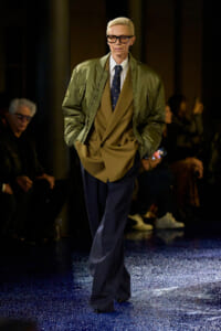 Model on a runway wearing an olive bomber over a tan suit with a white shirt, navy tie, and dark blue trousers, gloves optional.