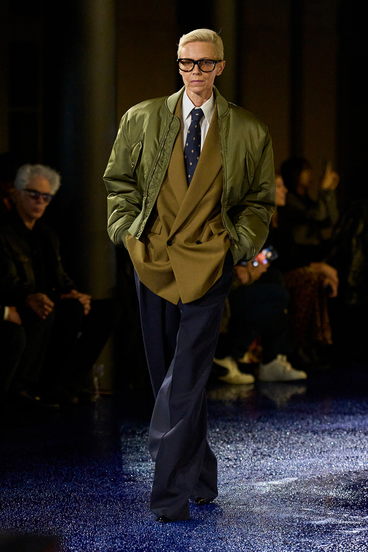 Model on a runway wearing an olive bomber over a tan suit with a white shirt, navy tie, and dark blue trousers, gloves optional.