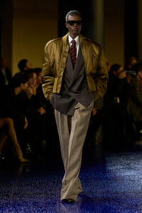 Model walking a runway in a gold-brown bomber jacket over a layered gray suit with a burgundy striped tie and dark sunglasses.
