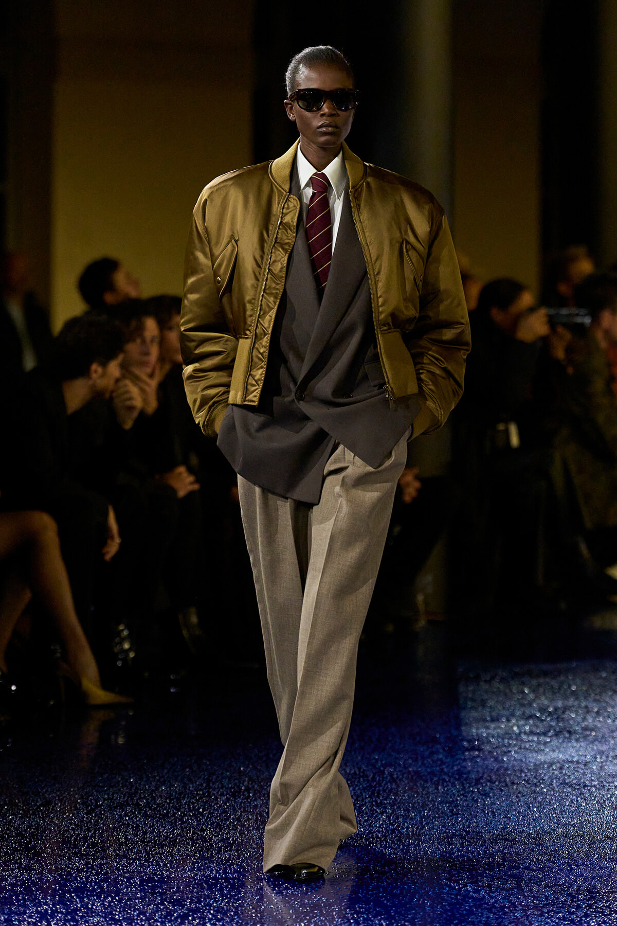 Model walking a runway in a gold-brown bomber jacket over a layered gray suit with a burgundy striped tie and dark sunglasses.
