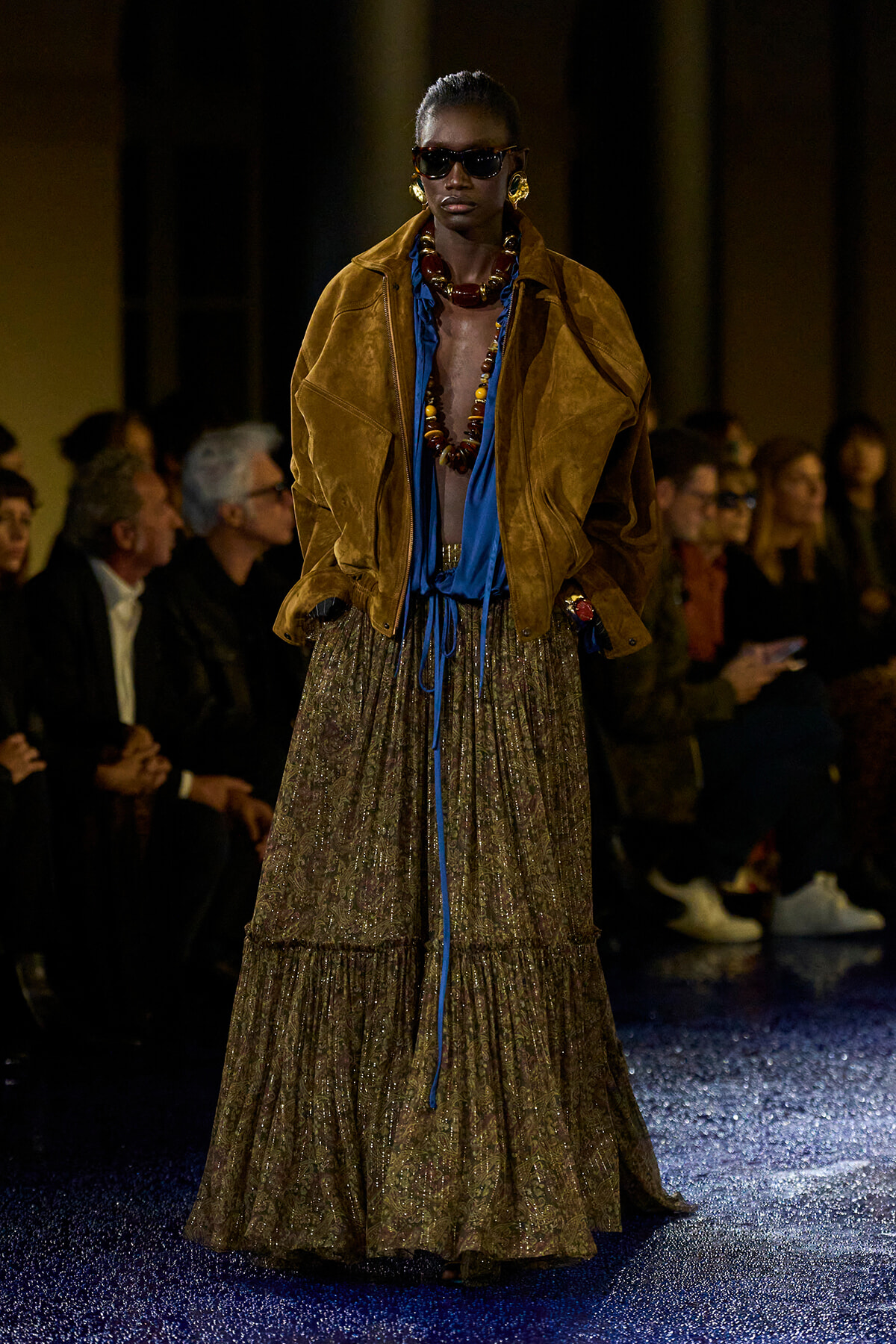 Model on runway wearing oversized brown suede jacket over a blue blouse, layered beaded necklace, and a long patterned skirt; dark sunglasses and bold jewelry.