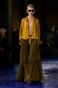 Model walks the runway in a mustard satin blouse and olive tiered maxi skirt, wearing dark sunglasses and bold earrings.