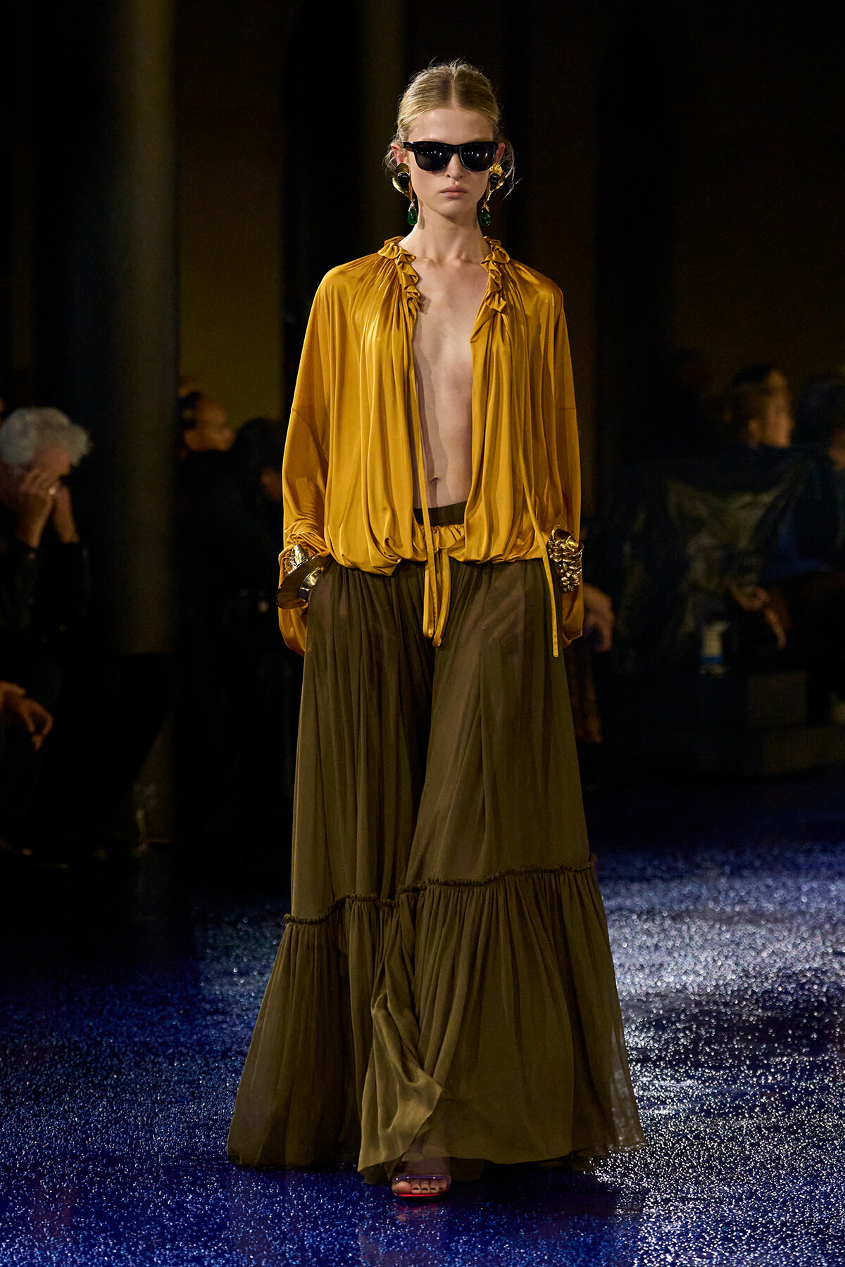 Model walks the runway in a mustard satin blouse and olive tiered maxi skirt, wearing dark sunglasses and bold earrings.