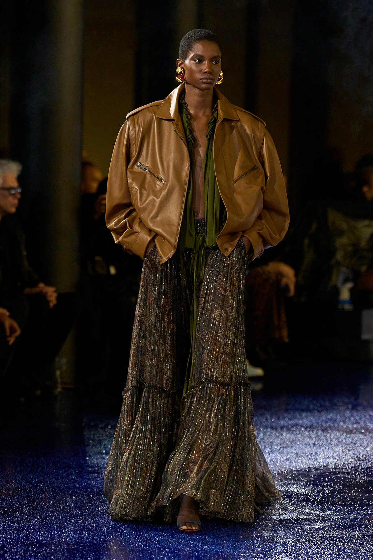 Model on runway wearing a tan oversized leather jacket over a green blouse and a long metallic skirt with glittery patterns and open sandals.