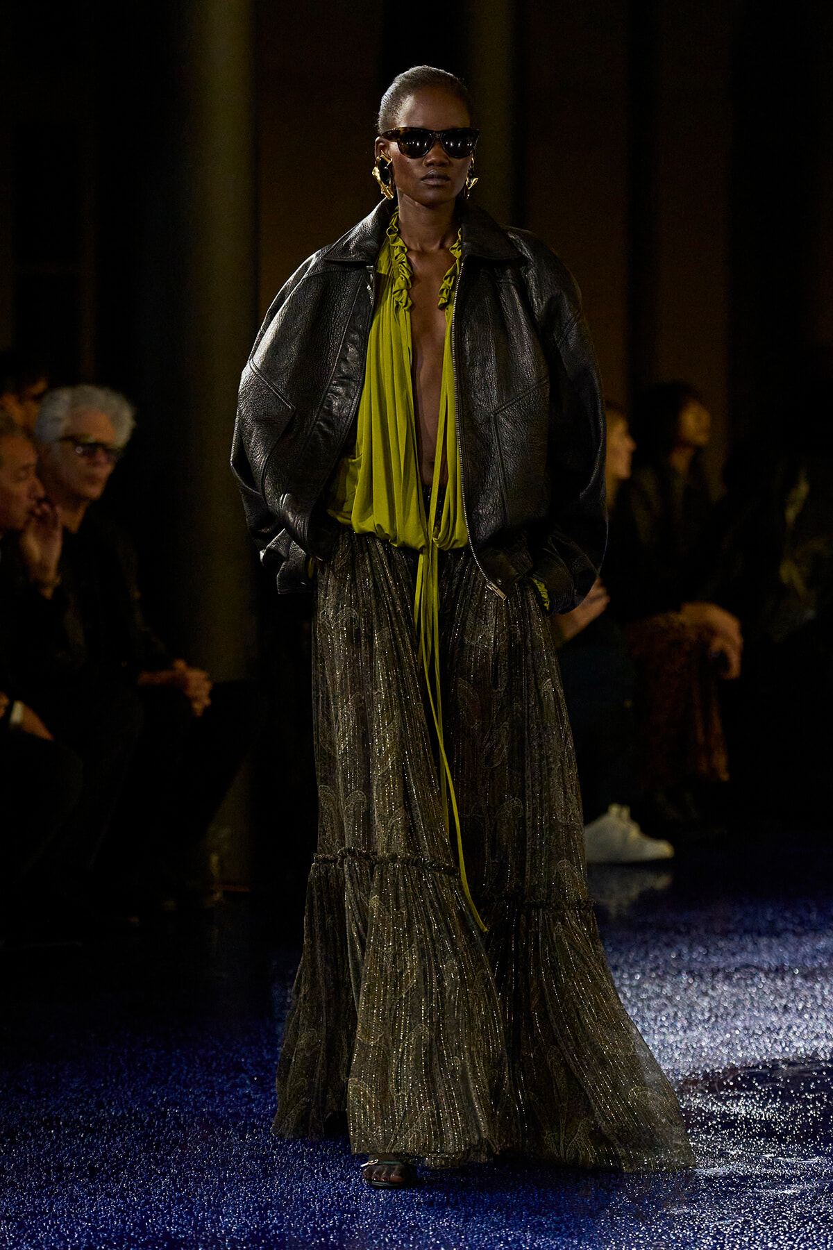 Runway model in an oversized black leather jacket over a lime green blouse and a shimmering, tiered maxi skirt with gold jewelry and dark sunglasses.