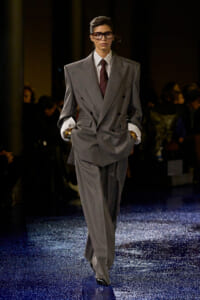 Model walking a fashion runway in a oversized gray double-breasted suit with striped shirt and burgundy tie, hands in pockets, wearing glasses
