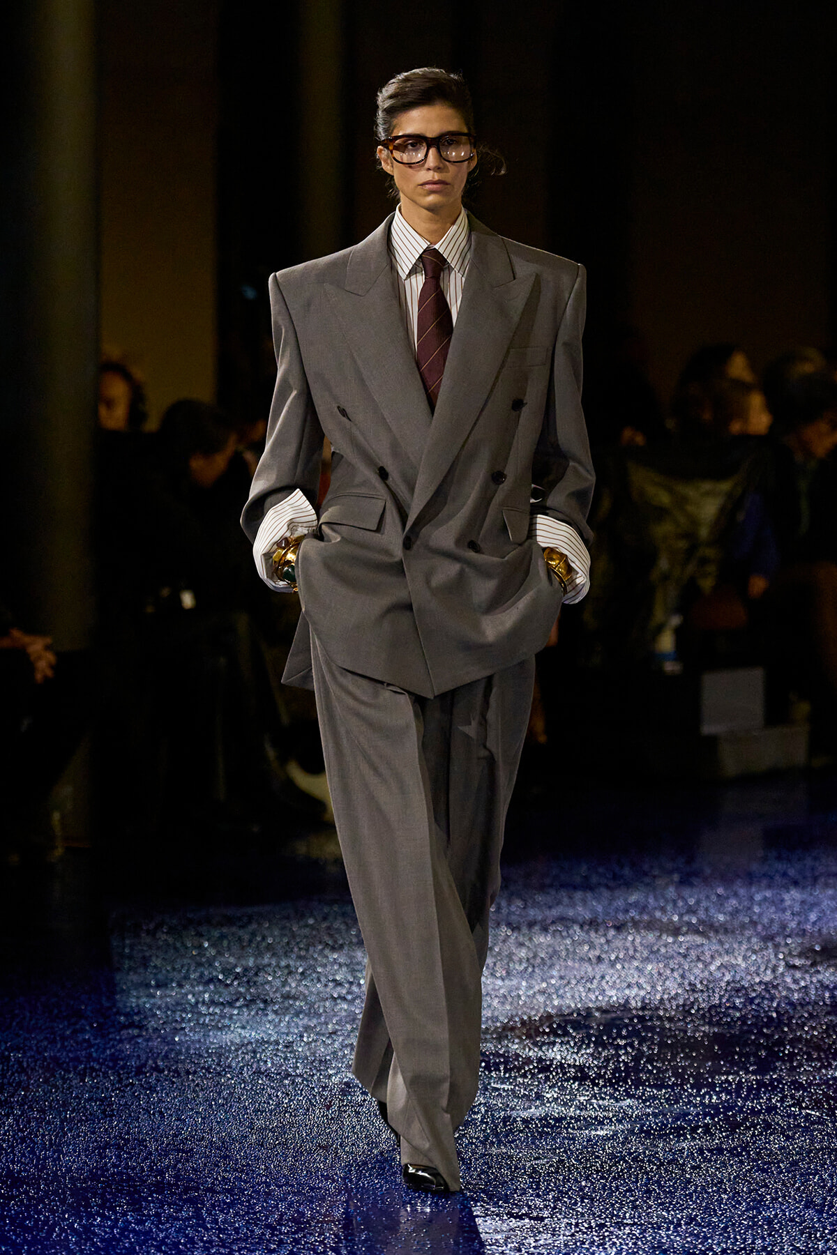 Model walking a fashion runway in a oversized gray double-breasted suit with striped shirt and burgundy tie, hands in pockets, wearing glasses
