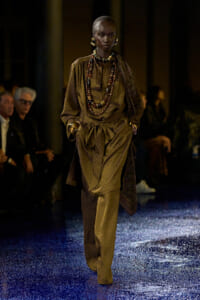 Model walking a fashion runway in a brown patterned draped suit with layered beaded necklaces and large earrings; blue glittery floor beneath.