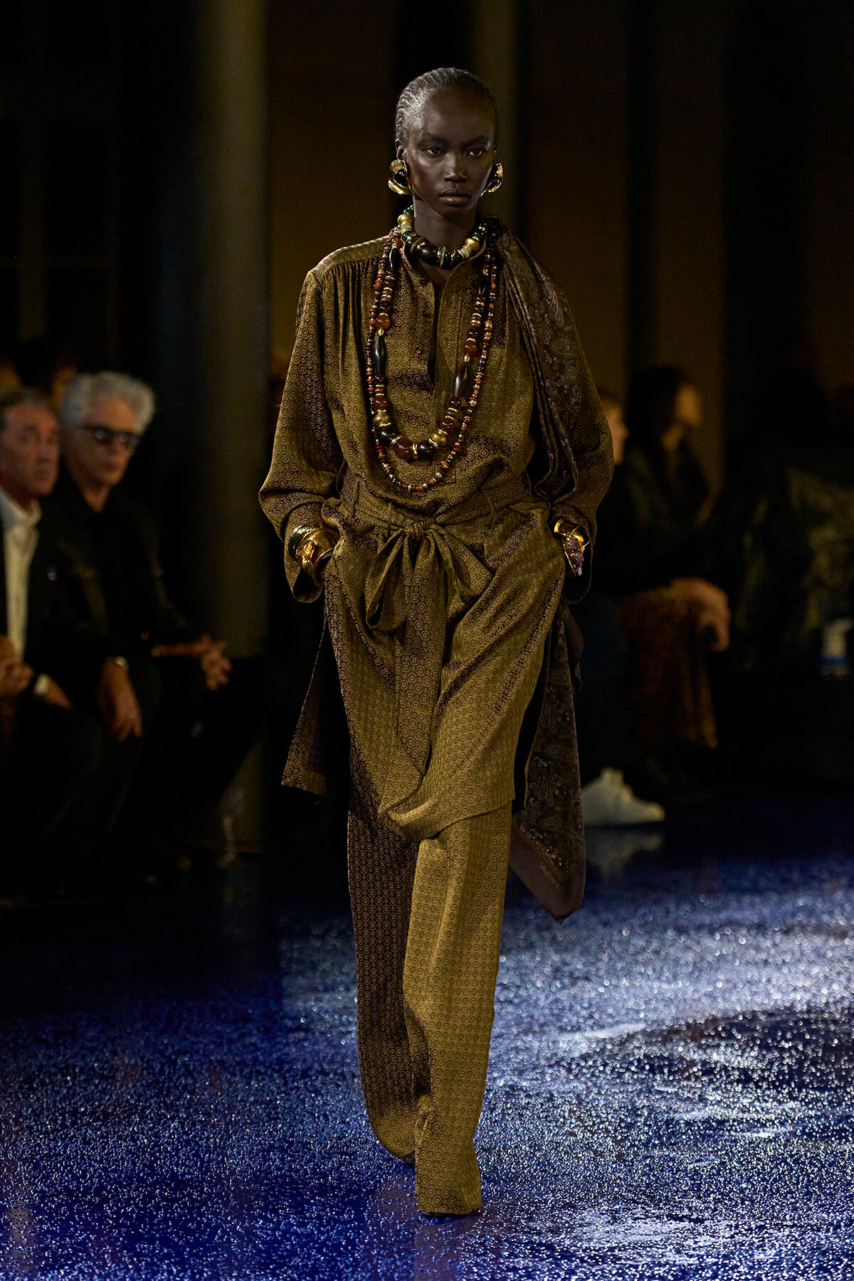Model walking a fashion runway in a brown patterned draped suit with layered beaded necklaces and large earrings; blue glittery floor beneath.