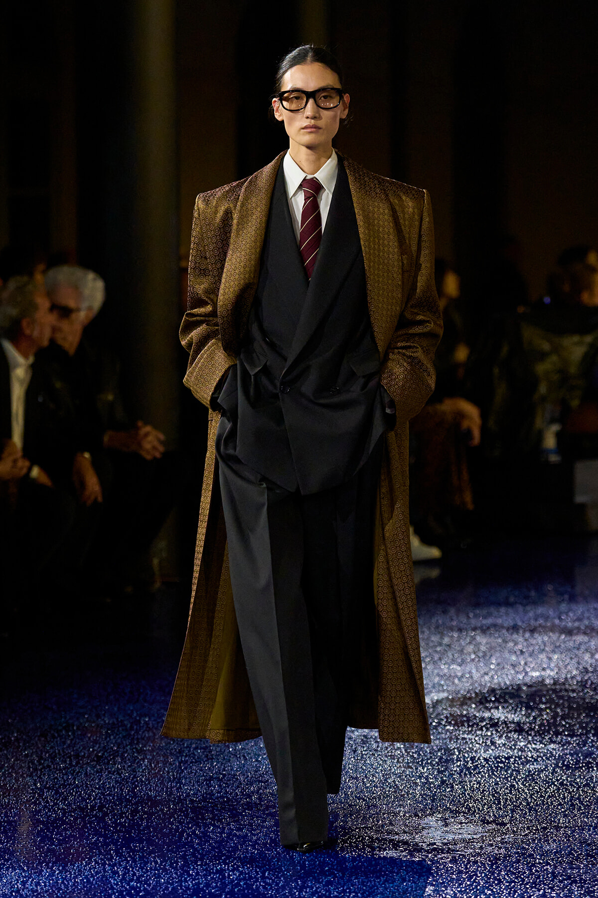 Model on a runway wearing a brown patterned long coat over a black draped suit, white shirt, maroon striped tie, and oversized glasses.