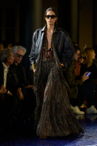 Model walking the runway in a dark leather jacket over a shimmering tiered gown, oversized sunglasses, and bold beaded necklaces.