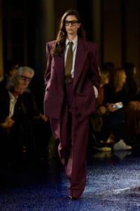 Model walks the runway in a burgundy oversized suit with a striped shirt and patterned tie, wearing dark glasses; audience in the background.