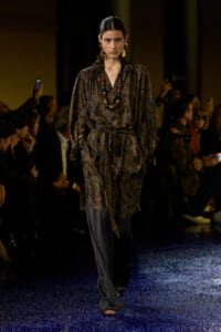 Model walking the runway in a dark paisley wrap dress with a long beaded necklace and bold gold earrings, on a glittery blue carpet.