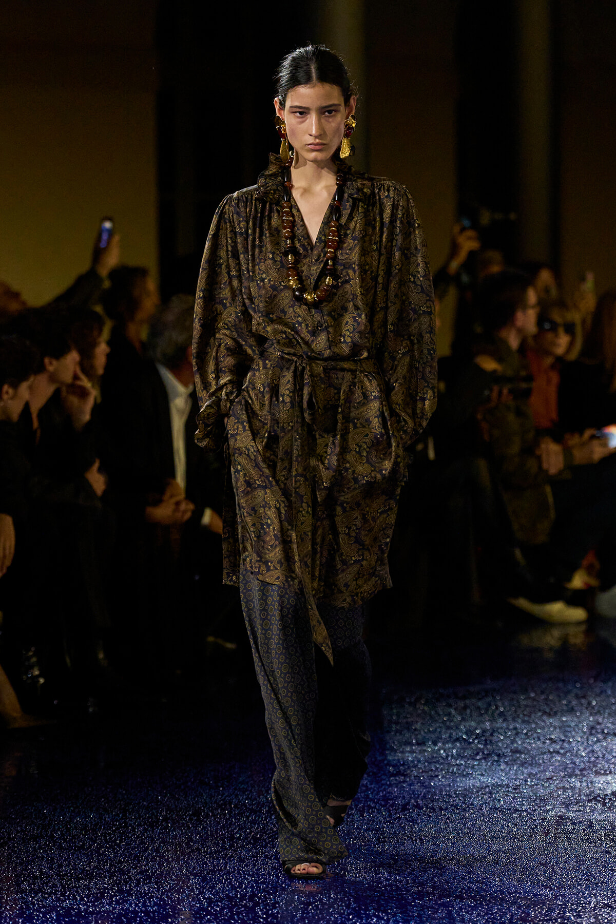 Model walking the runway in a dark paisley wrap dress with a long beaded necklace and bold gold earrings, on a glittery blue carpet.