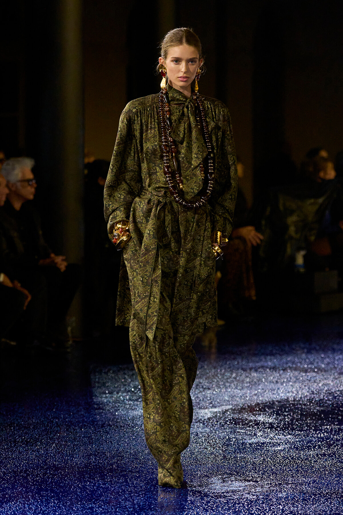 Model walking the runway in a green paisley suit with a bow-tied neck, layered beaded necklace and gold jewelry.