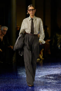 Model walking a runway in a striped button-down shirt, dark wide-leg trousers, and a black polka-dot tie, holding a jacket.