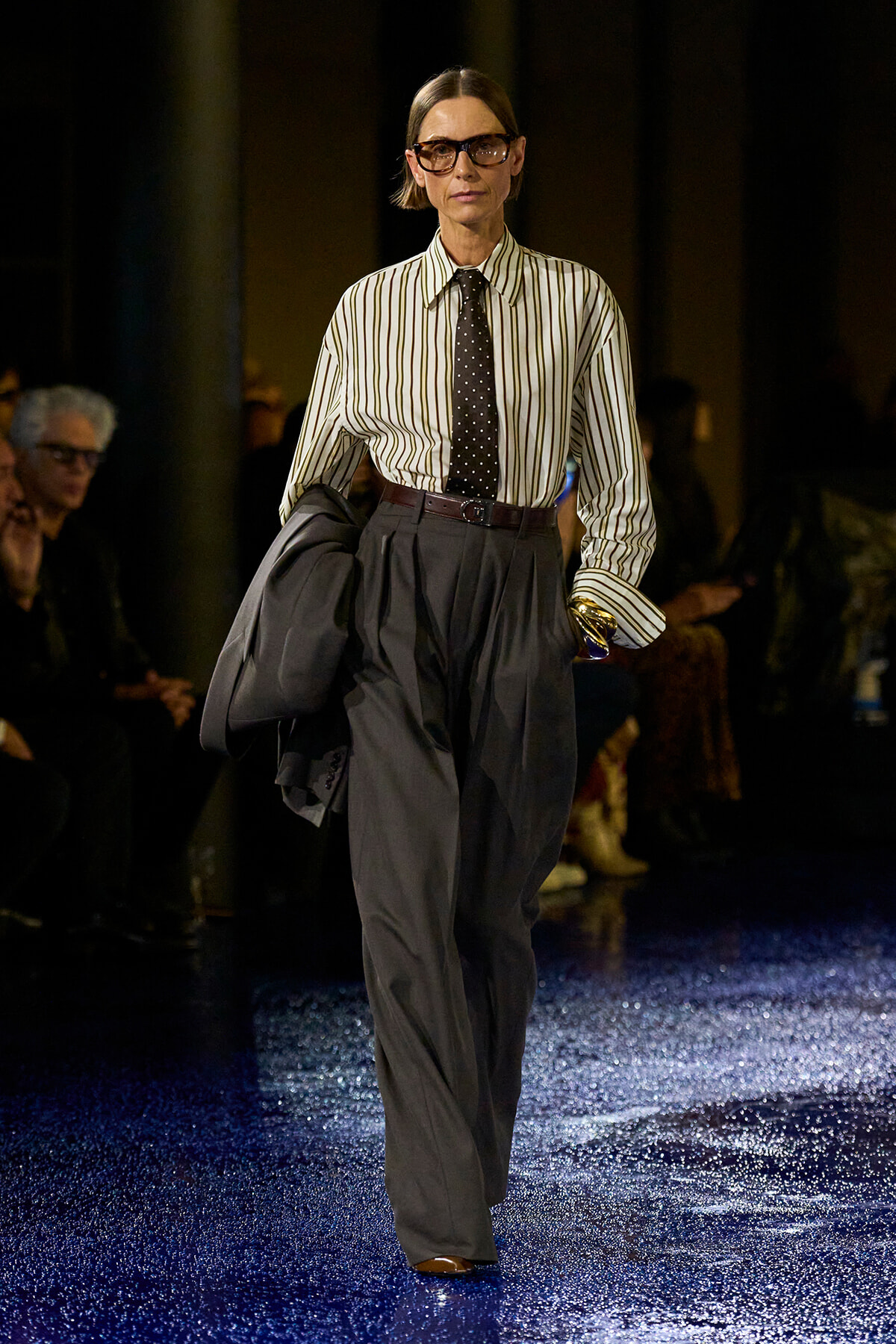 Model walking a runway in a striped button-down shirt, dark wide-leg trousers, and a black polka-dot tie, holding a jacket.