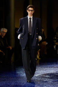 Male model walking a runway in an oversized navy double-breasted blazer, striped tie, white shirt, and wide-framed glasses.