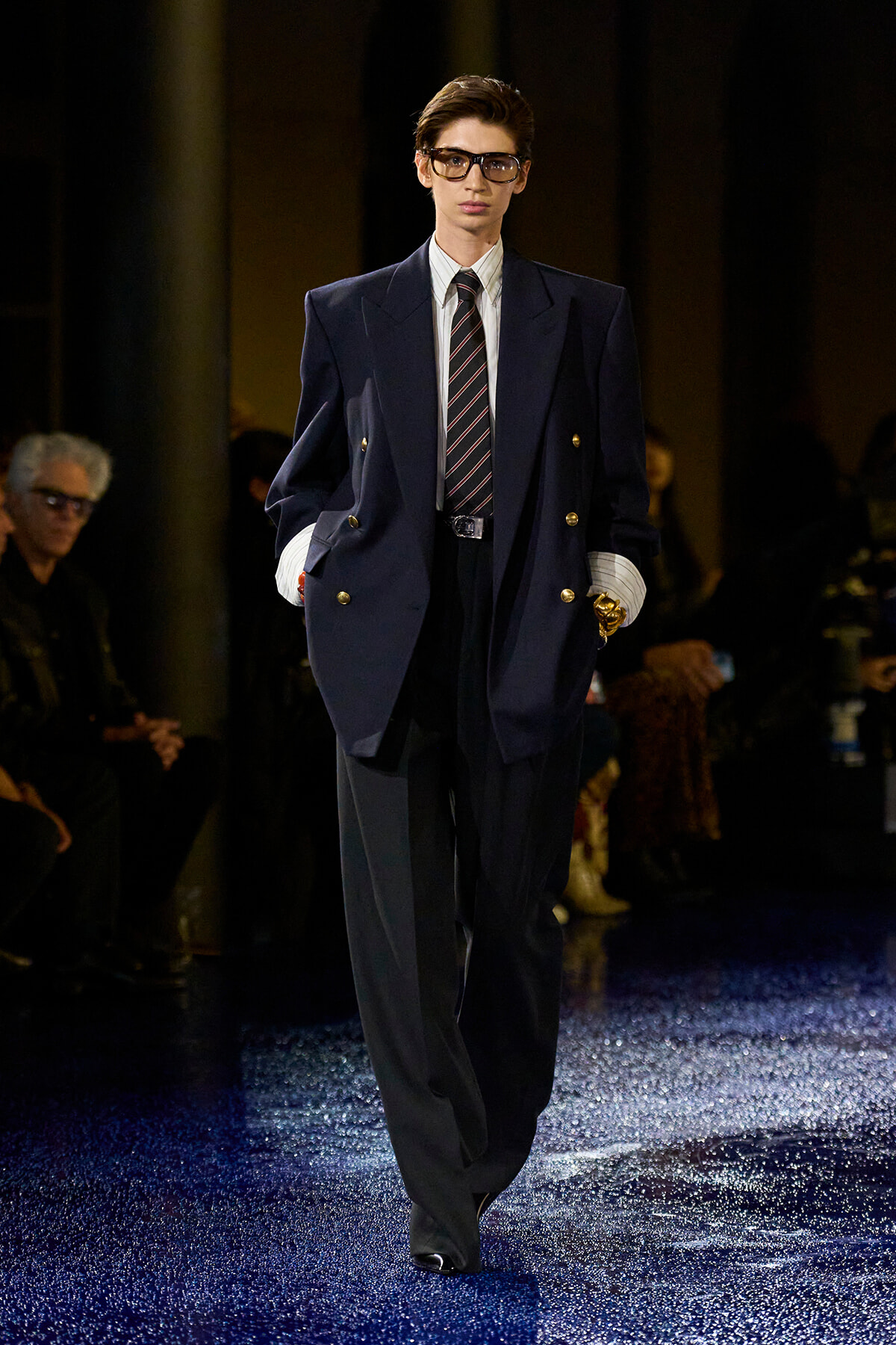 Male model walking a runway in an oversized navy double-breasted blazer, striped tie, white shirt, and wide-framed glasses.