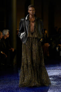 Model on a fashion runway wearing a black leather jacket, plunging metallic top, layered gold jewelry, and a long paisley-printed skirt rocketing down the catwalk.