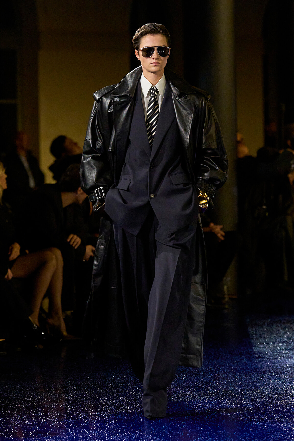 Male model in an oversized black leather trench over a navy suit with a striped tie and sunglasses, walking the runway on a blue-glittered catwalk.