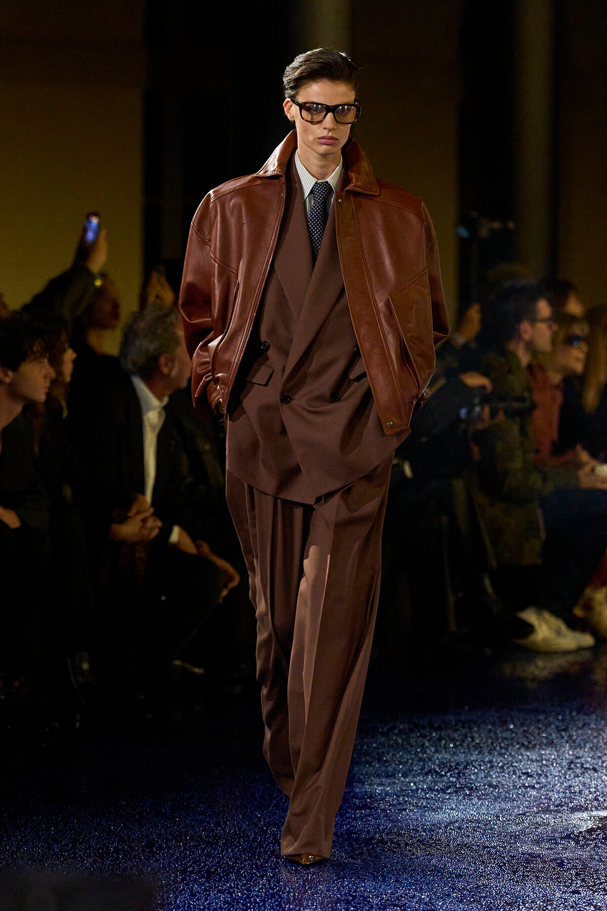 Male model on a runway wearing a brown leather jacket over a brown suit with a white shirt and dark tie, sunglasses on.