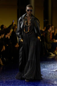 Model walking a runway in a long black dress and oversized black leather coat, layered beaded necklaces, gold cuffs, and dark sunglasses.