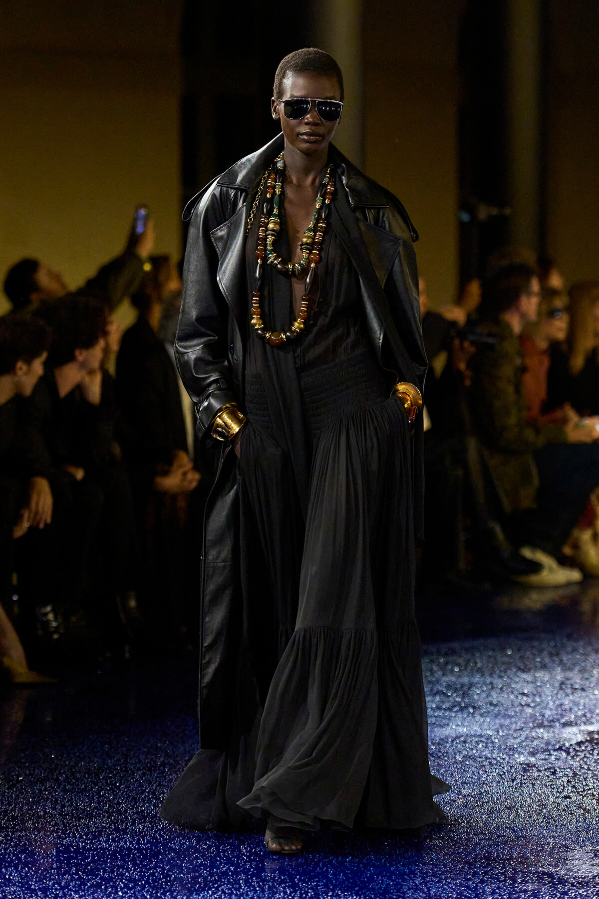 Model walking a runway in a long black dress and oversized black leather coat, layered beaded necklaces, gold cuffs, and dark sunglasses.