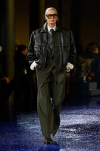 Model in an oversized black leather jacket over a dark green suit with a striped tie, wearing sunglasses on a glittery blue runway.