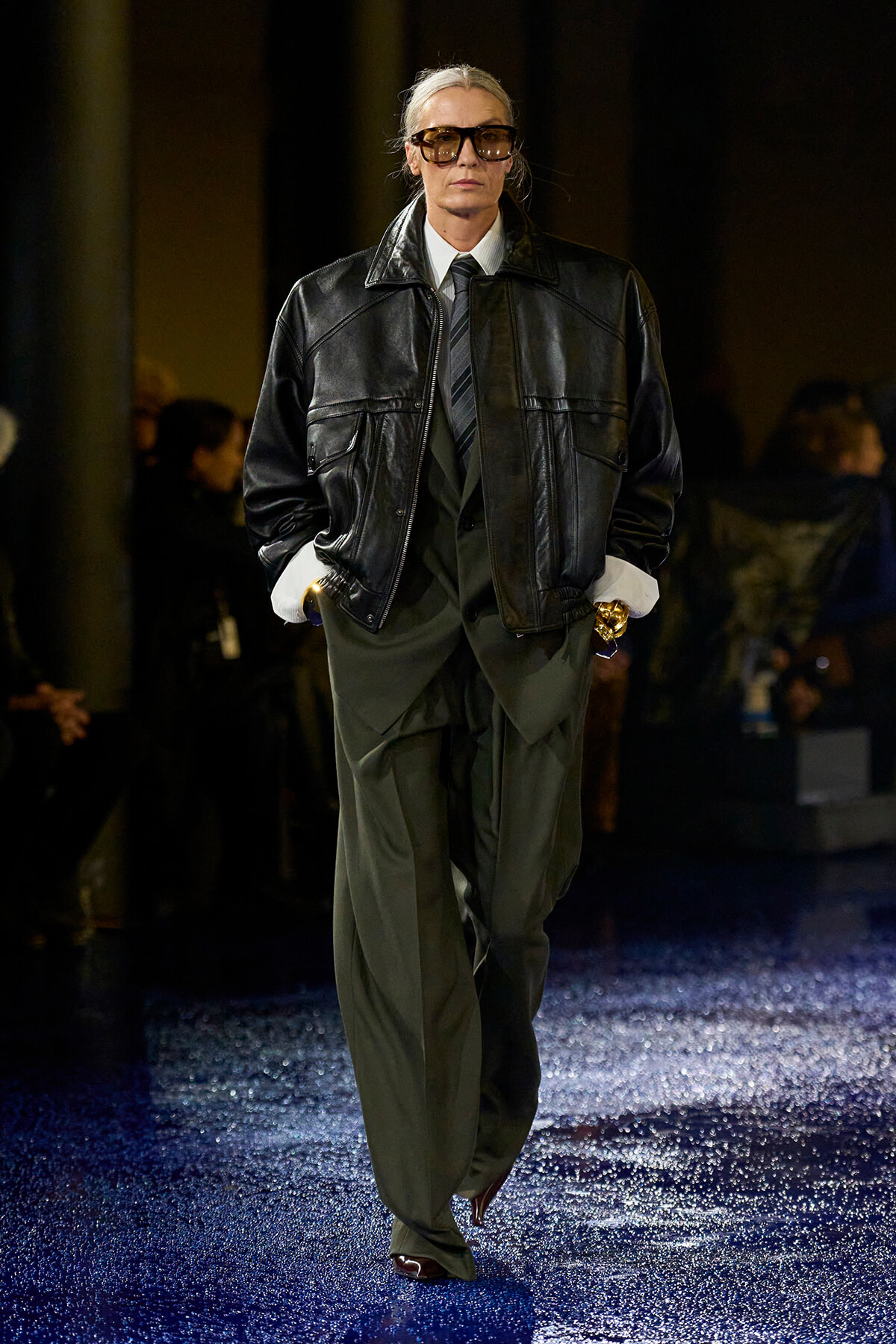 Model in an oversized black leather jacket over a dark green suit with a striped tie, wearing sunglasses on a glittery blue runway.
