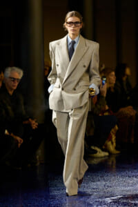 Model on the runway in an oversized light gray double-breasted suit with a blue shirt, dark tie, and glasses, walking confidently.