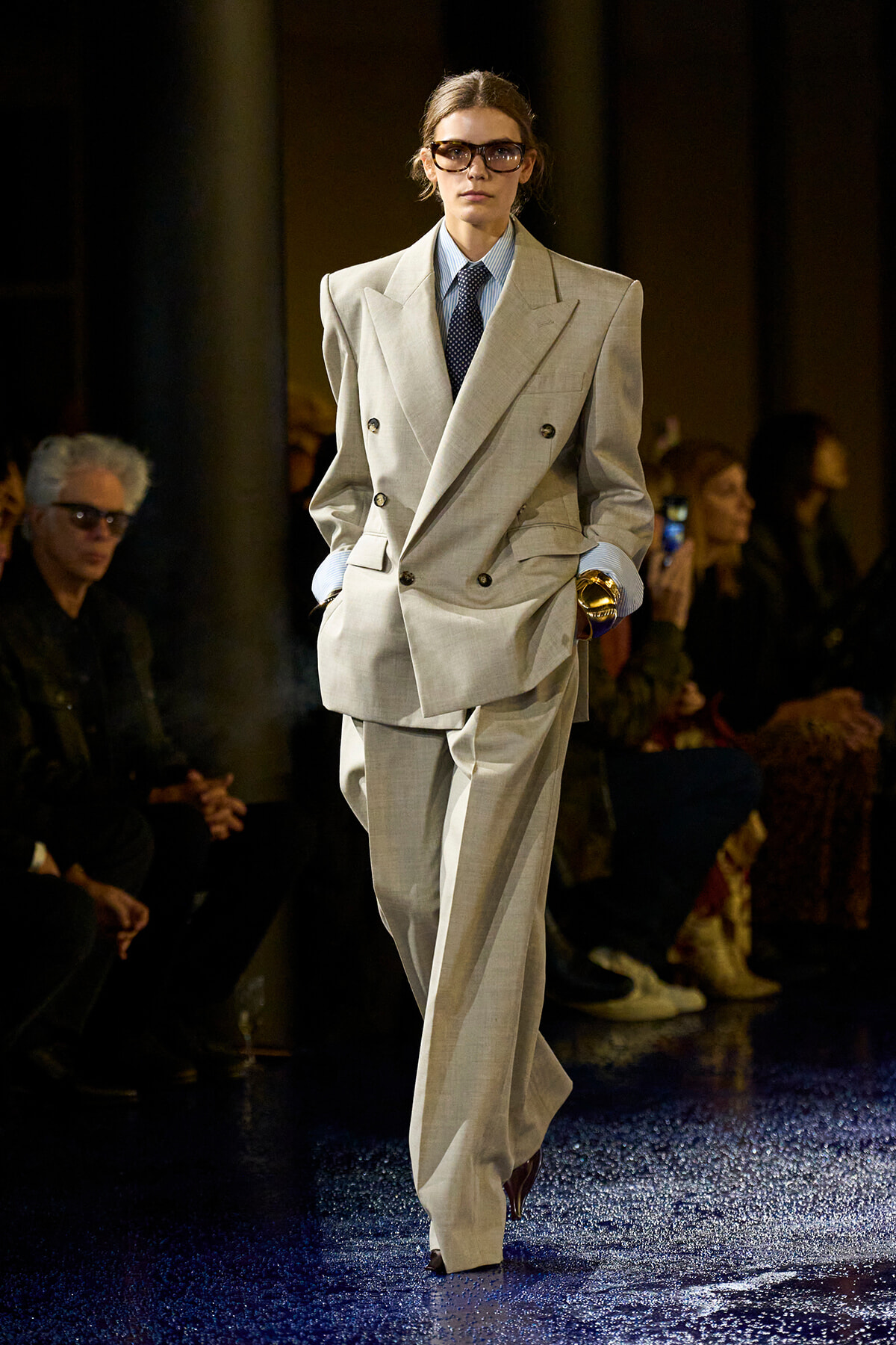 Model on the runway in an oversized light gray double-breasted suit with a blue shirt, dark tie, and glasses, walking confidently.