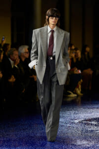 Model on the runway wearing an oversized gray suit with a striped shirt and burgundy tie, black glasses, audience in the background.