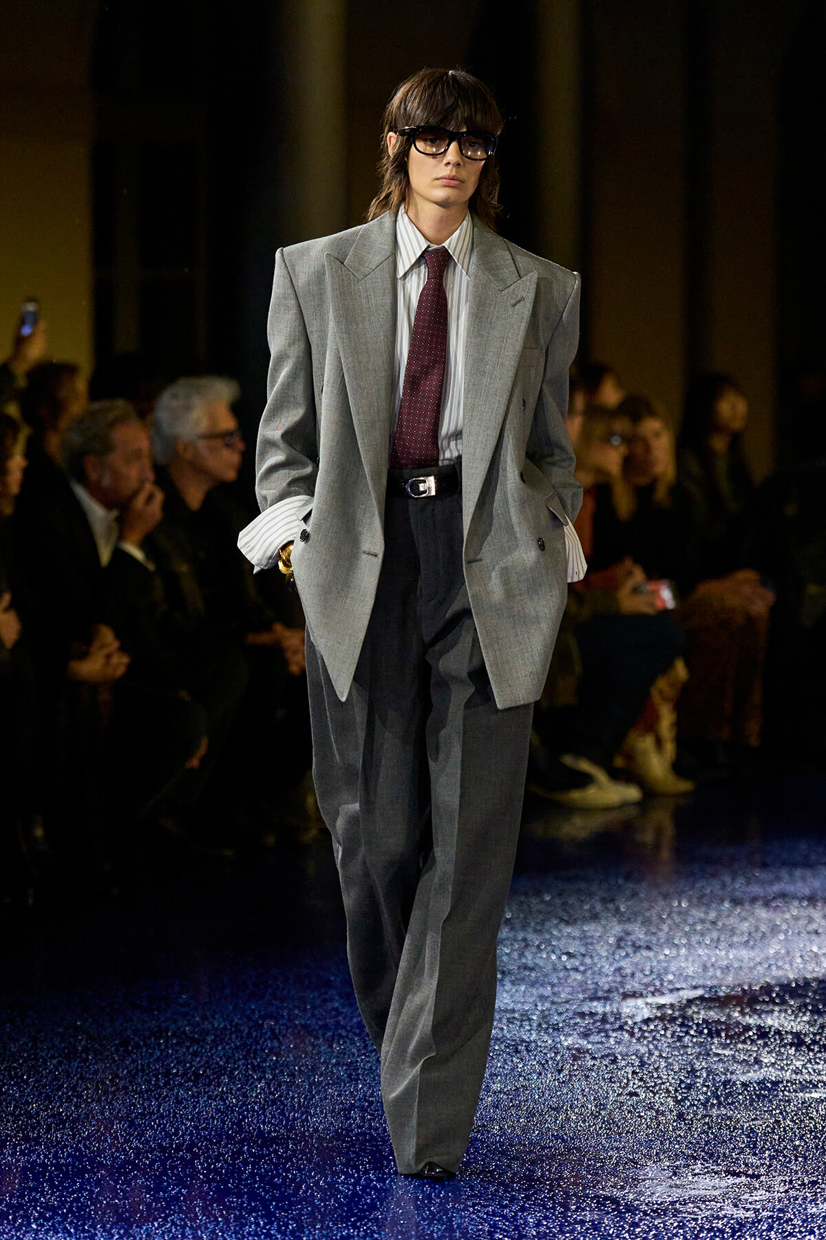 Model on the runway wearing an oversized gray suit with a striped shirt and burgundy tie, black glasses, audience in the background.