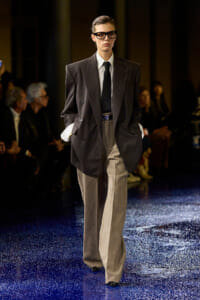 Model on a runway wearing an oversized dark blazer, white shirt, black tie, and wide beige trousers with a blue belt.