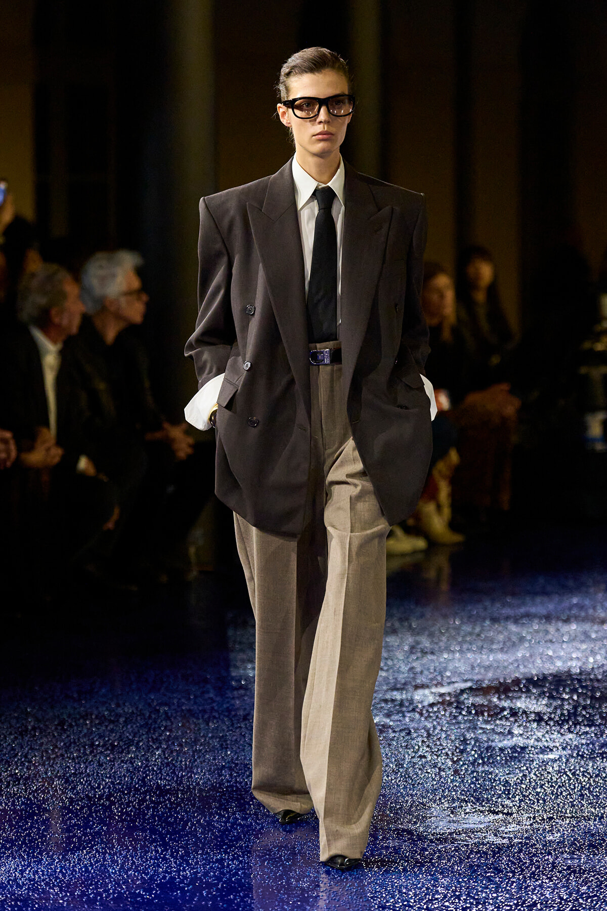 Model on a runway wearing an oversized dark blazer, white shirt, black tie, and wide beige trousers with a blue belt.