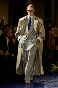 Model walking a runway in a light gray double-breasted suit with an oversized beige trench coat and blue tie; audience watches in dim backdrop.