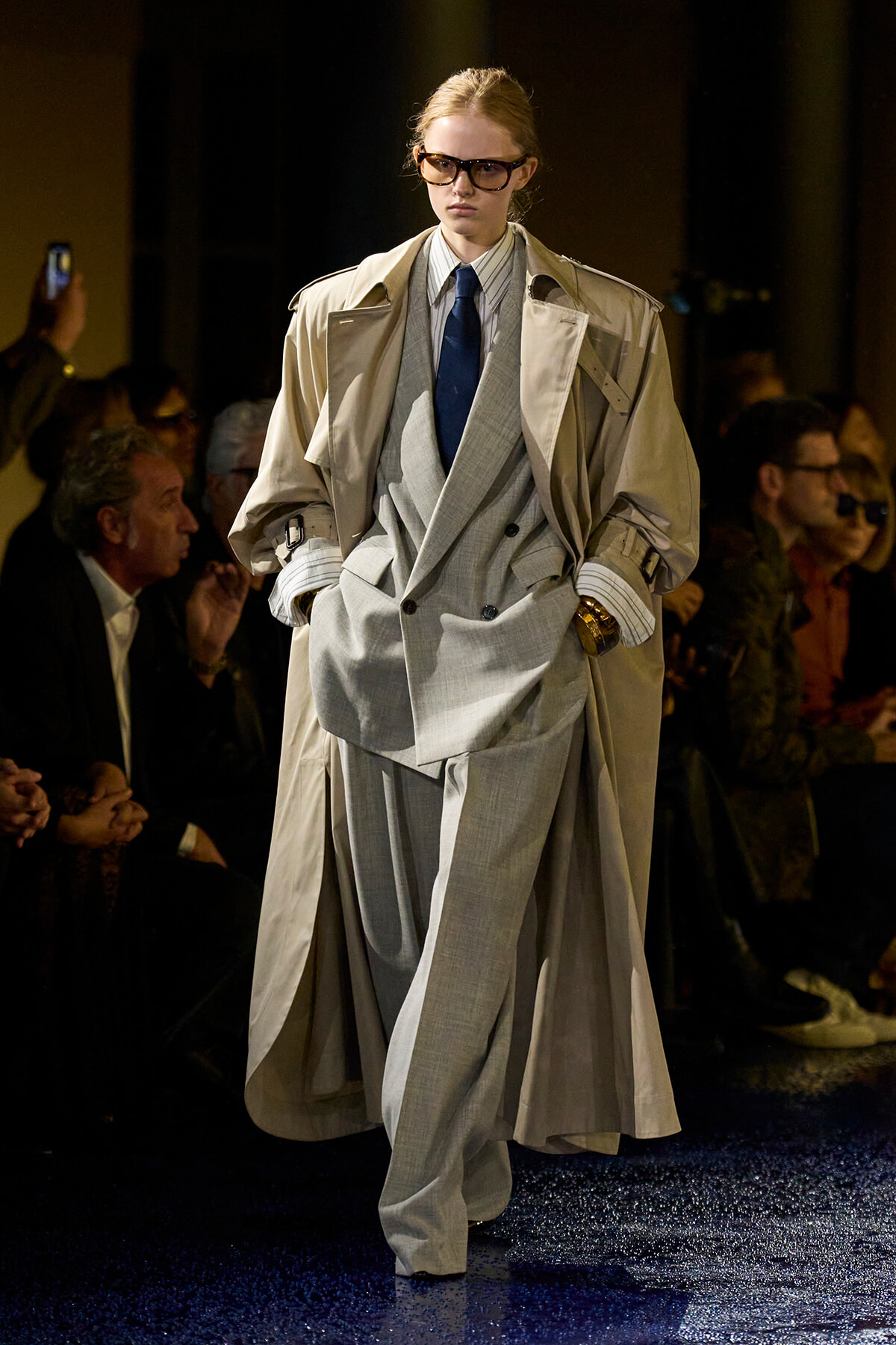 Model walking a runway in a light gray double-breasted suit with an oversized beige trench coat and blue tie; audience watches in dim backdrop.