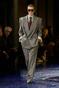 Model on a runway wearing an oversized light-gray double-breasted suit with a burgundy tie and dark sunglasses.