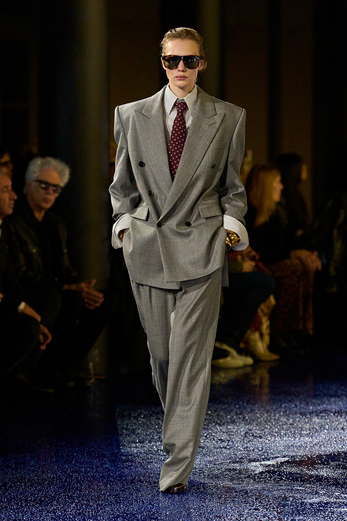 Model on a runway wearing an oversized light-gray double-breasted suit with a burgundy tie and dark sunglasses.