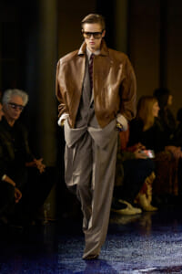 Male runway model wearing a brown leather bomber jacket over a gray pinstripe suit with a burgundy tie and sunglasses, walking the catwalk.