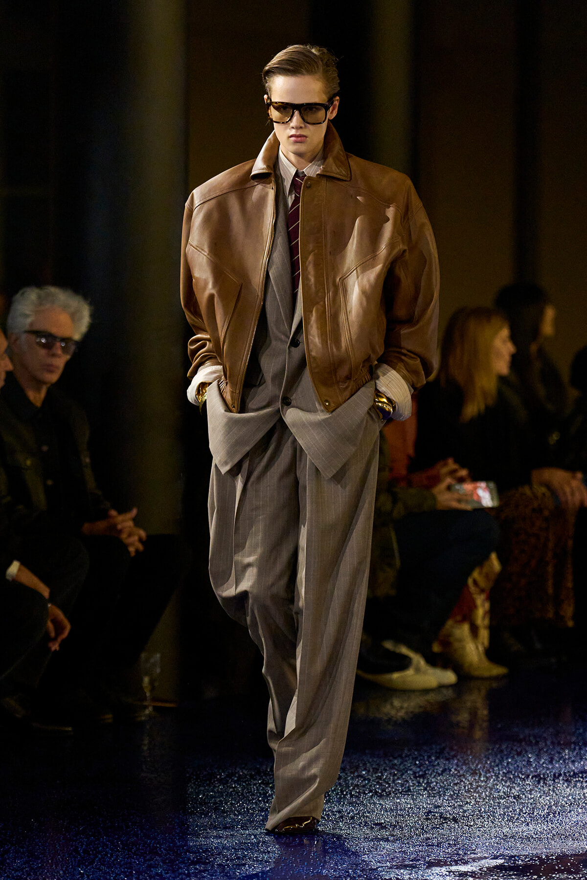 Male runway model wearing a brown leather bomber jacket over a gray pinstripe suit with a burgundy tie and sunglasses, walking the catwalk.